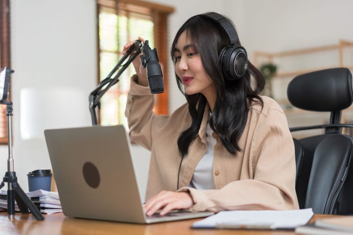 female podcaster wearing headphones speaking into microphone on laptop preparing to record a podcast remotely
