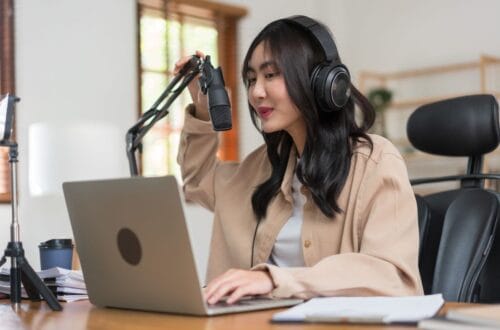 female podcaster wearing headphones speaking into microphone on laptop preparing to record a podcast remotely
