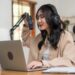 female podcaster wearing headphones speaking into microphone on laptop preparing to record a podcast remotely